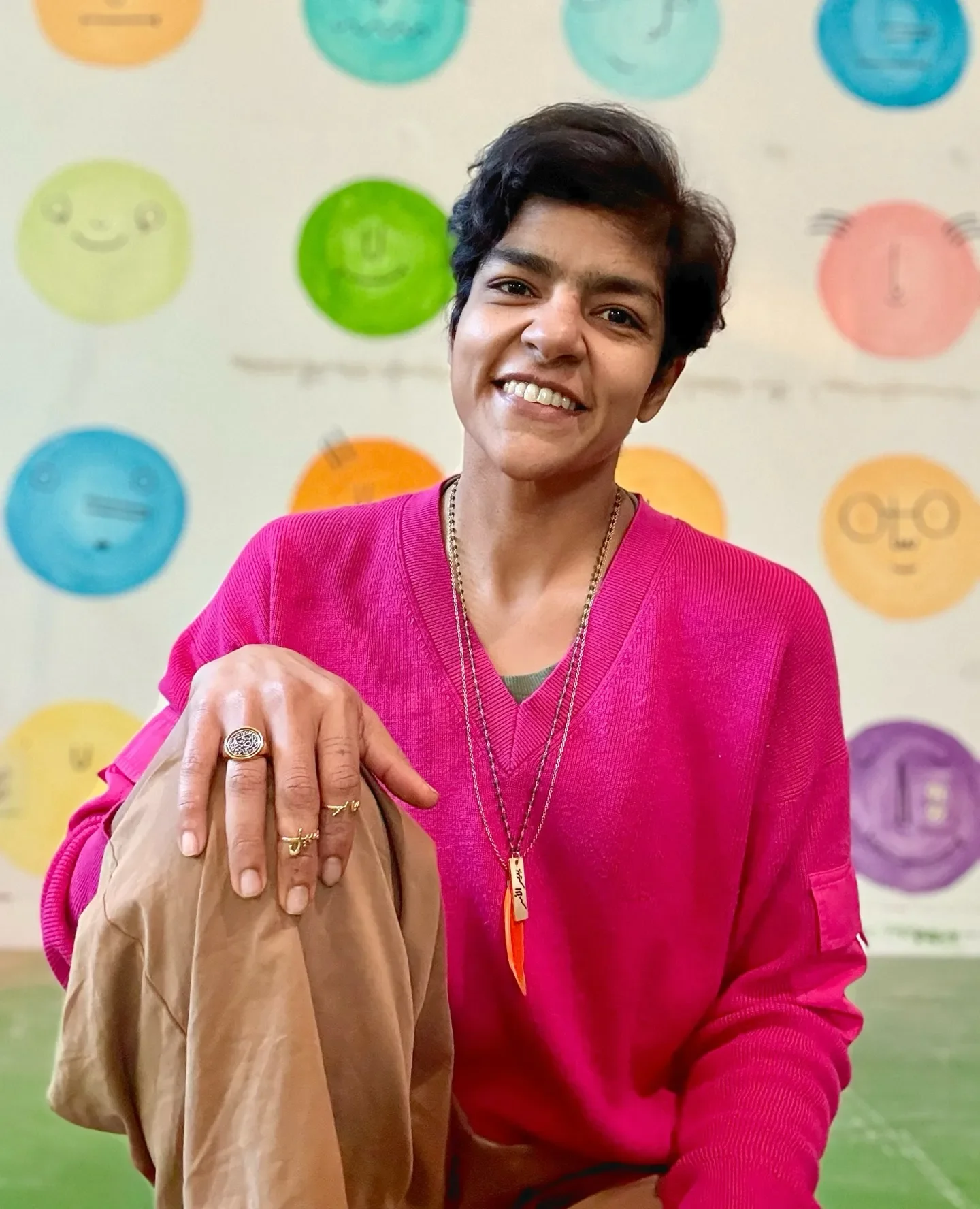 Areej Khan Founder of Foster Within, smiling, sitting in Sesh pose, wearing a bright pink sweater and tan pants. Background features colorful, feelings faces on the wall behind her at Foster Within.