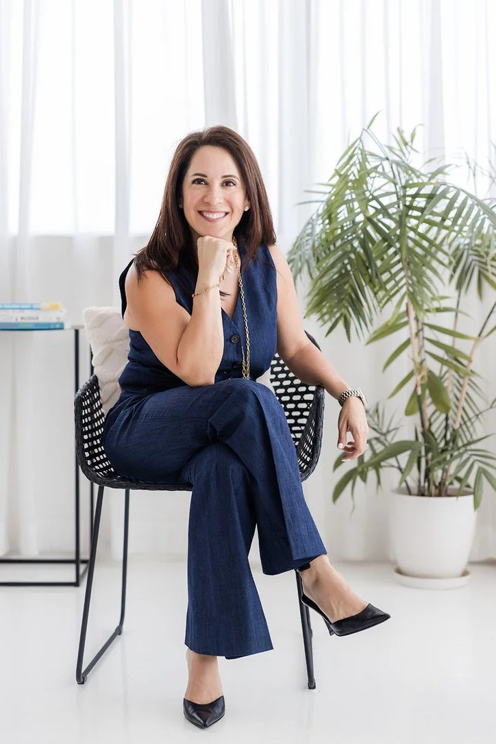 A woman in a sleeveless blue jumpsuit sitting on a black modern chair, smiling at the camera, with a large green potted plant and white curtains in the background.