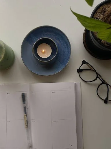 A desk with a blue candle on a matching blue saucer, a black potted plant with green leaves, a pair of black glasses, a gray pen, and an open planner or notebook with blank pages.