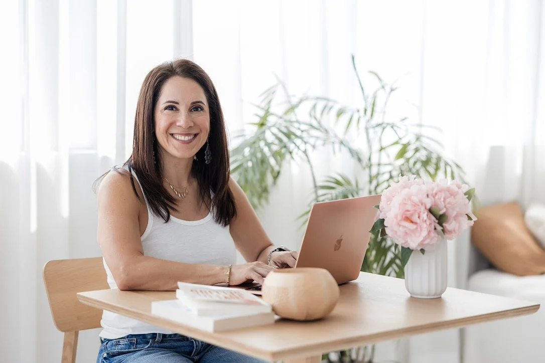 A woman smiling while working on a pink laptop at a wooden table with a vase of pink flowers and a book, in a bright room with white curtains and green plants.