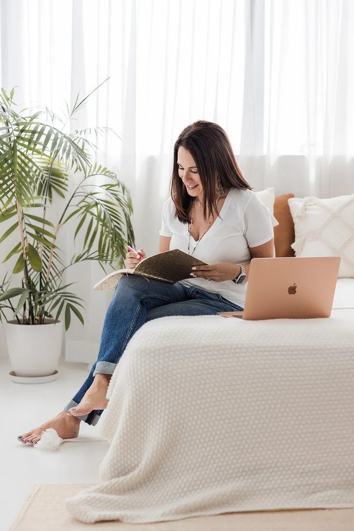 A woman sits on a bed, reading a notebook with a pink pen, with a laptop open beside her, in a bright room with sheer white curtains and a large indoor plant.