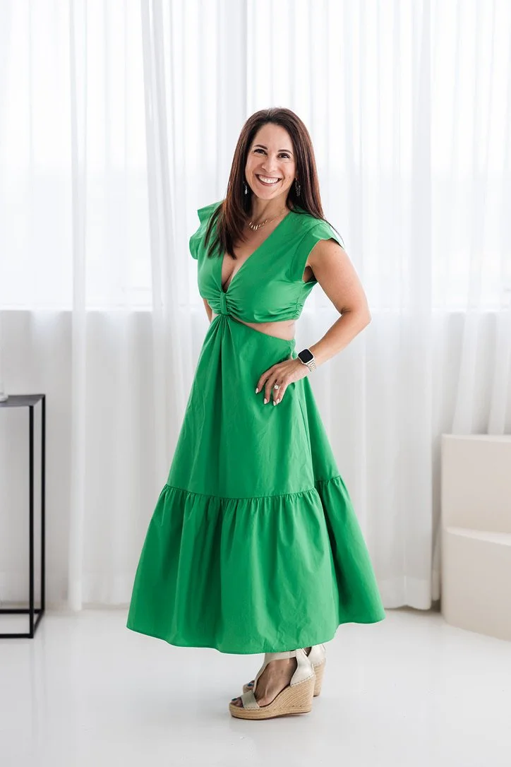 Woman in a bright green dress and beige wedge sandals standing indoors with white curtains and a small black table in the background.