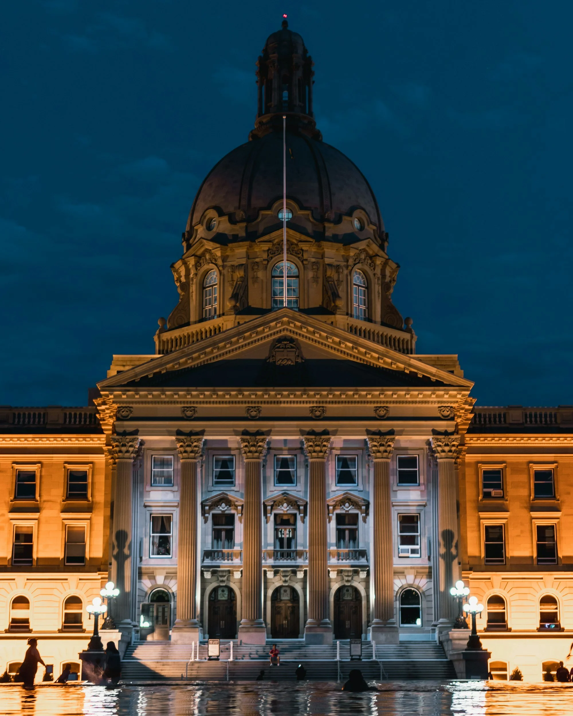 The Alberta government building illuminated at night, reflecting in the water in front of it.