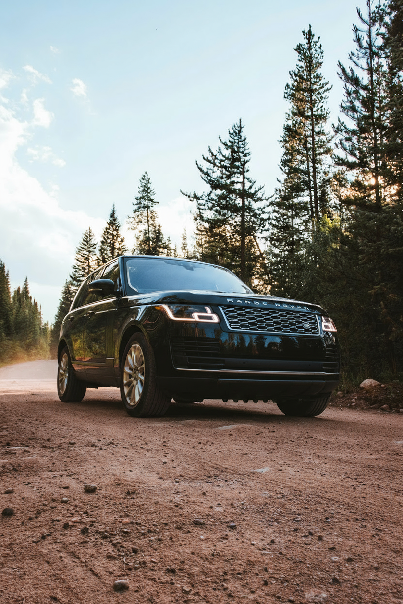 A black Range Rover driving on a dirt road through a forest with tall pine trees and a partly cloudy sky.