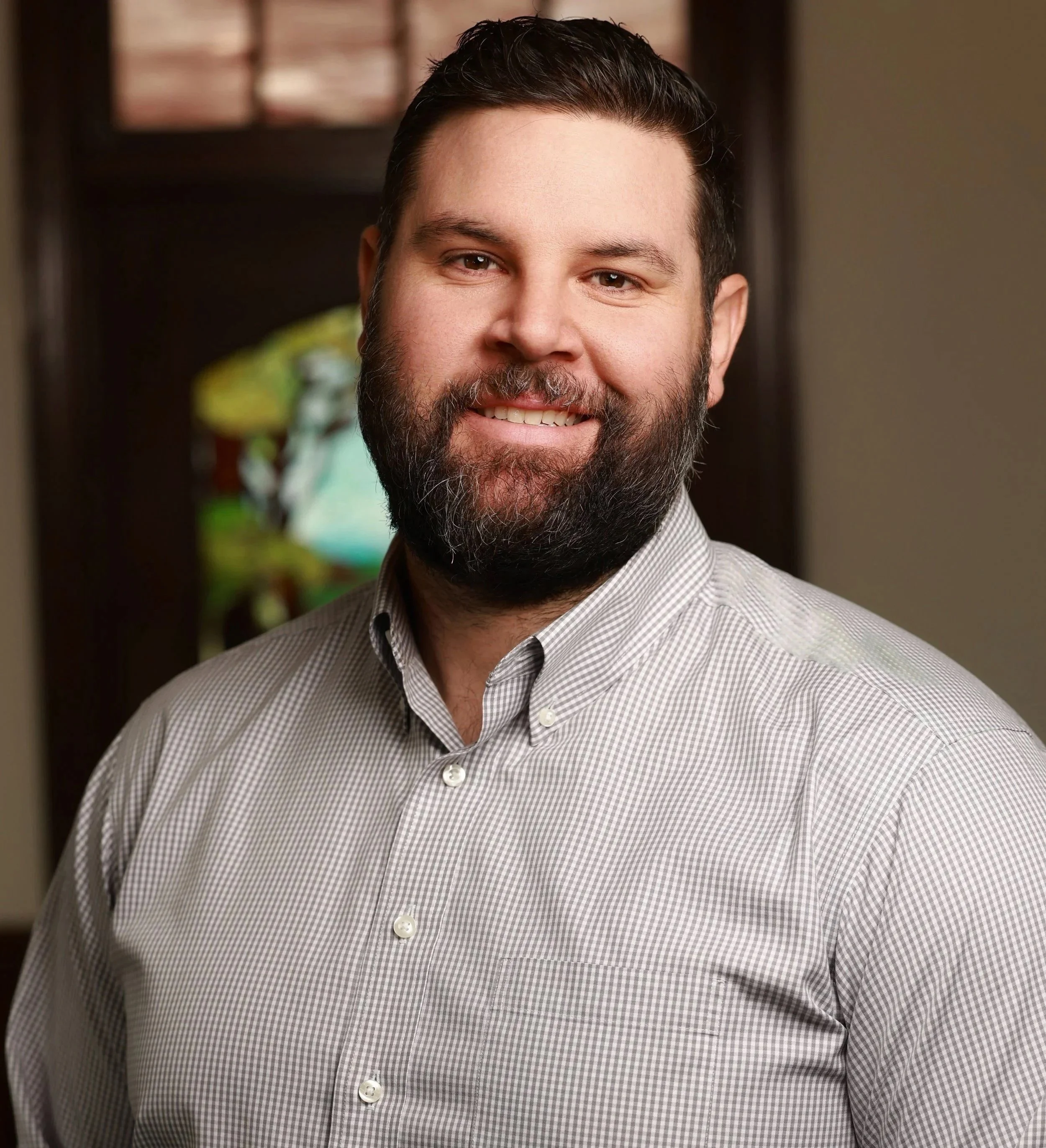 A smiling man with dark hair and a beard, wearing a light-colored checkered dress shirt, standing indoors with a blurred background.