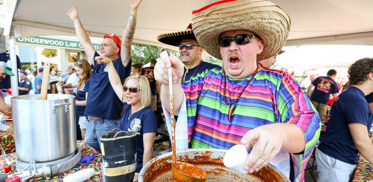 Man in colorful striped shirt and wide sombrero pouring sauce from a container, wearing sunglasses, at food festival with others in the background.