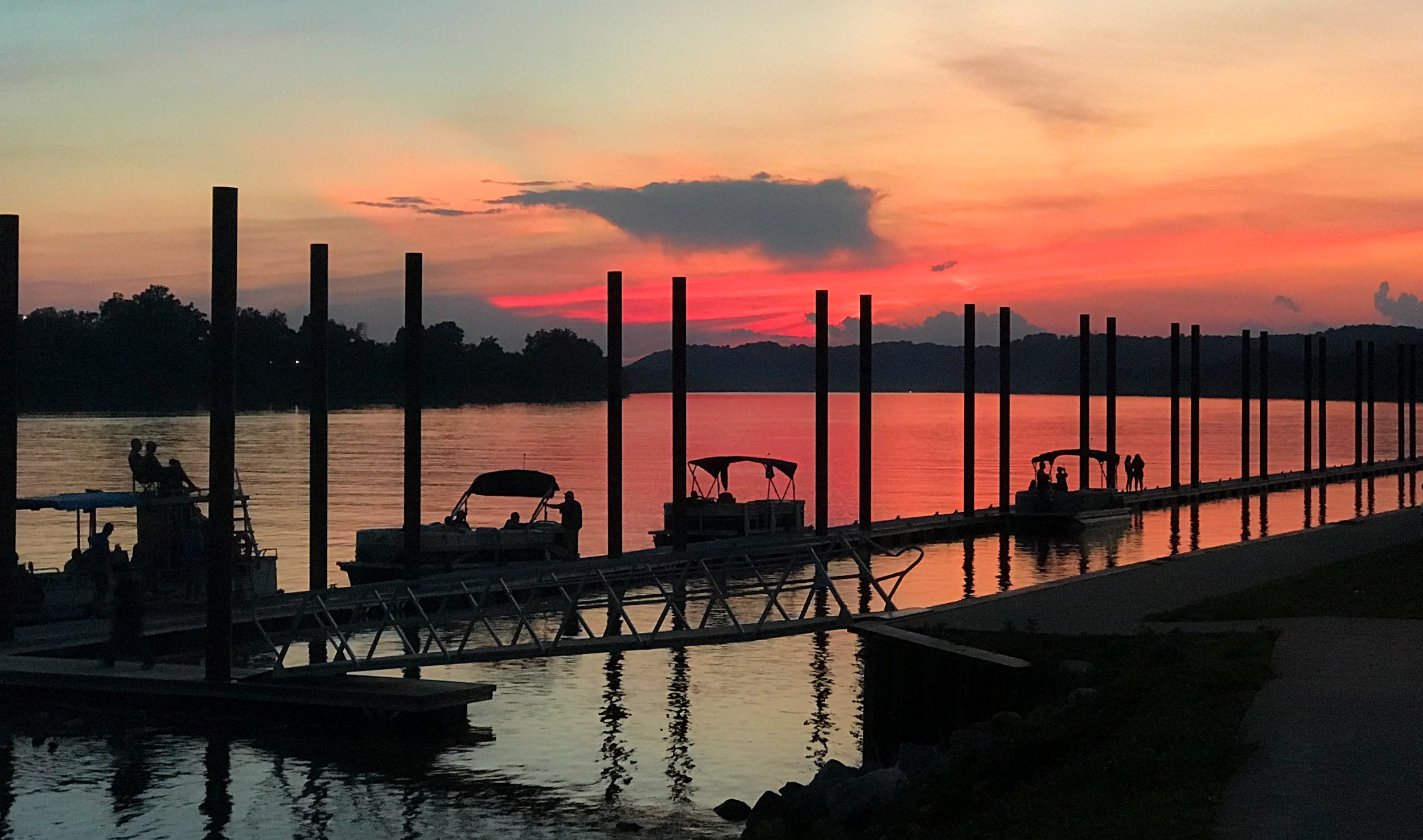 A photo of a boat dock at sunset with boats moored along a pier and people walking or sitting, silhouettes against a colorful sky with orange, pink, and purple hues reflected on the water.