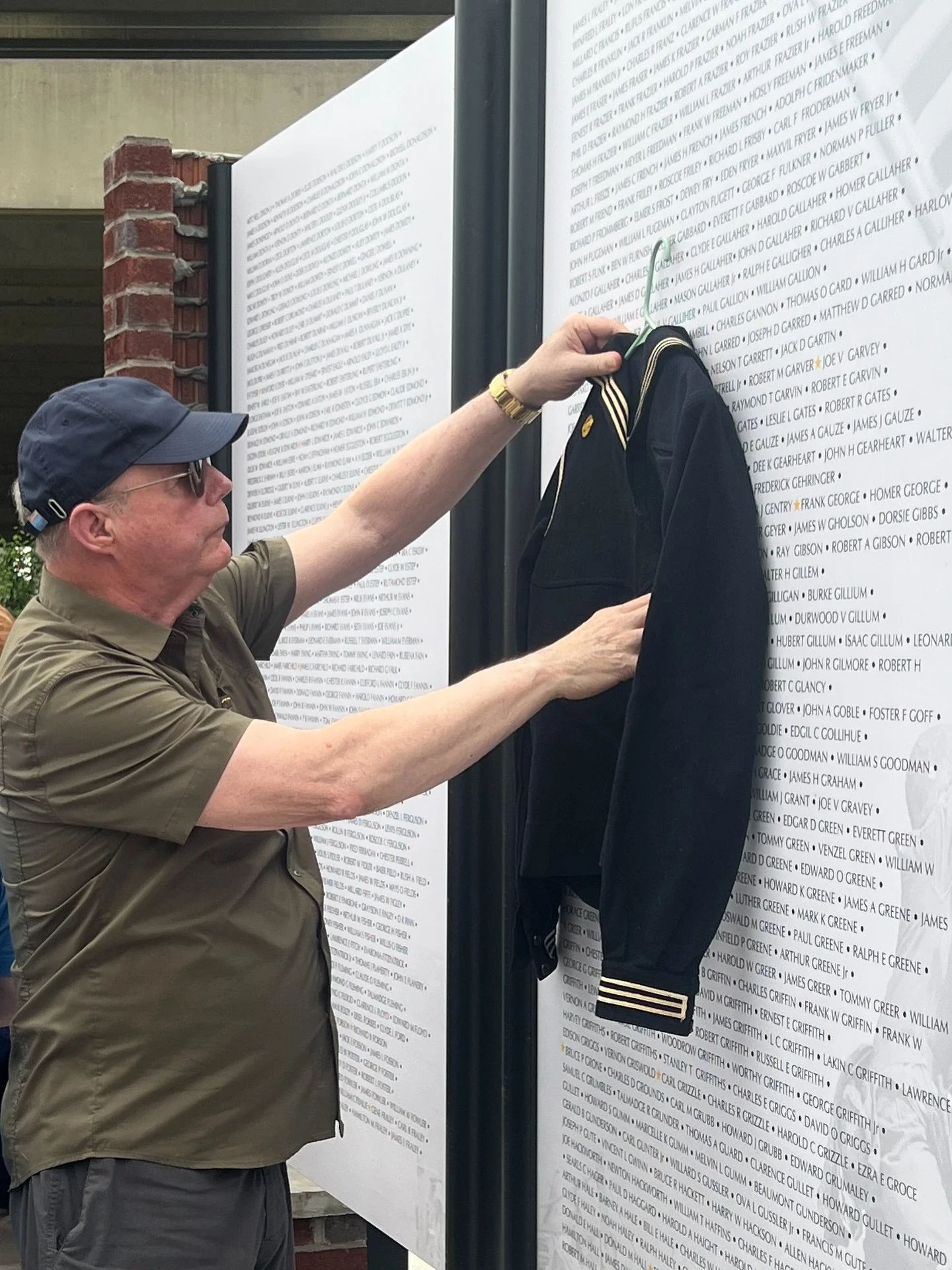 A man wearing a khaki shirt, glasses, and a navy cap is hanging a navy jacket with gold stripes on the sleeves on a memorial wall with numerous names inscribed on it.