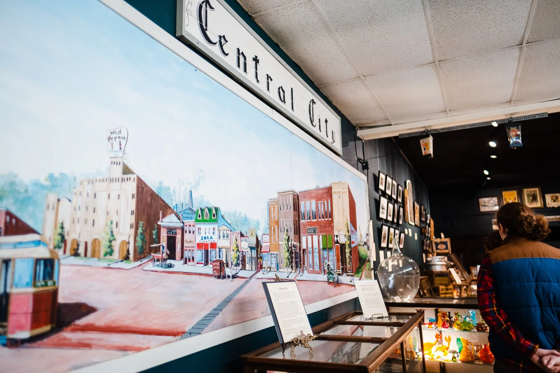 A large painting or mural of a street scene with buildings, shops, and a trolley, displayed on a wall in an art gallery or shop. A person is seen observing the artwork.