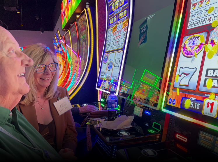 Two women smiling and playing slot machines in a colorful casino with bright lights and digital screens.