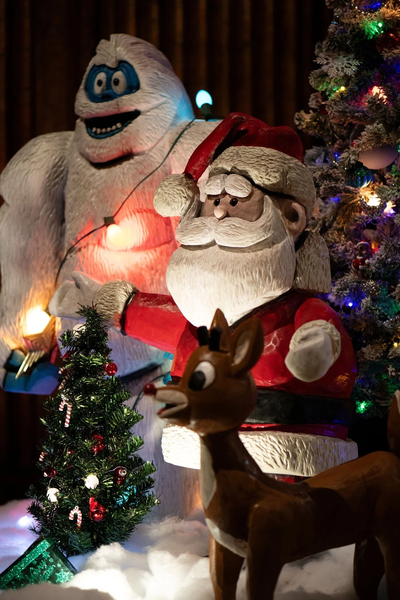 Decorative holiday display with a Santa Claus figure, a reindeer figure, a small decorated Christmas tree, and a large snowman figure, all illuminated with colorful Christmas lights.