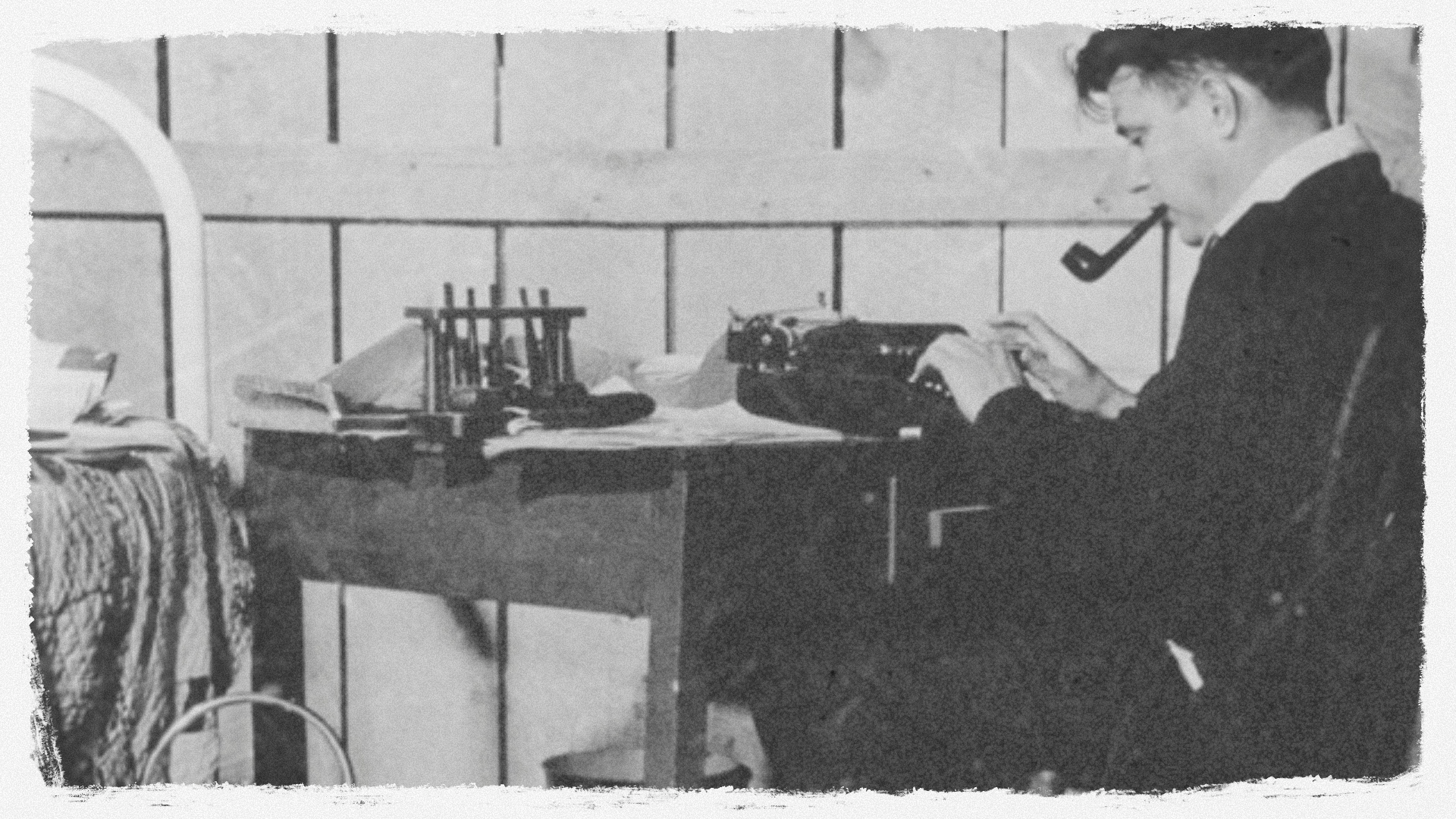 A person sitting at a desk typing on a typewriter, with a pipe in their mouth, surrounded by various objects including shoes and papers, in an indoor setting with a tiled wall background.
