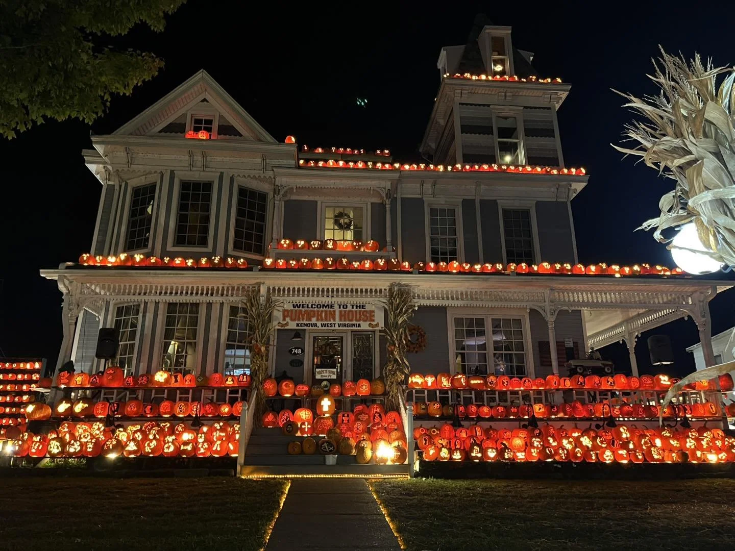 A large Victorian-style house decorated with numerous glowing orange and red pumpkins for Halloween, with a sign that reads 'Welcome to the Pumpkin House' in Kenova, West Virginia. The house has pumpkins on the porch and roof, with a dark night sky in the background.
