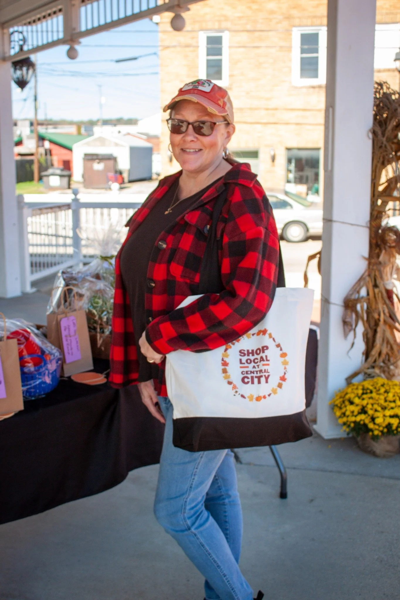A smiling woman wearing sunglasses, a red plaid jacket, and a beige cap, carrying a tote bag that says 'Shop Local at Central City'. She is standing outside near a table with gift bags, with a porch railing and a street with buildings in the background.