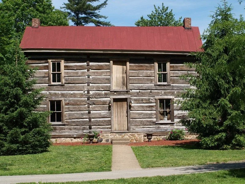 A rustic wooden house with a red metal roof, surrounded by green trees and a manicured lawn, with a concrete pathway leading to the front door.