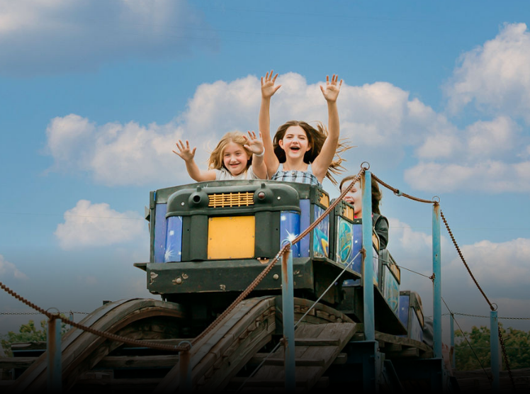 Three children with joyful expressions waving from a ride at an amusement park on a partly cloudy day.