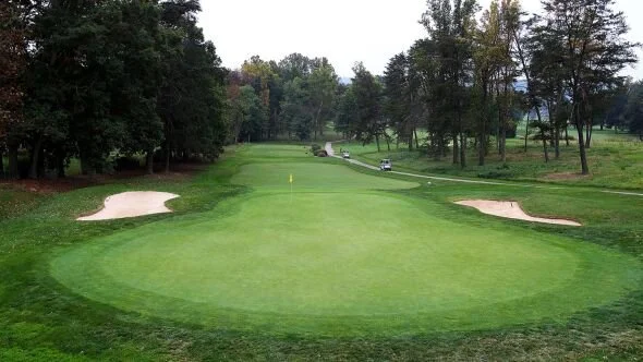 A golf course putting green with sand bunkers on either side, surrounded by trees and a small cart path in the background.