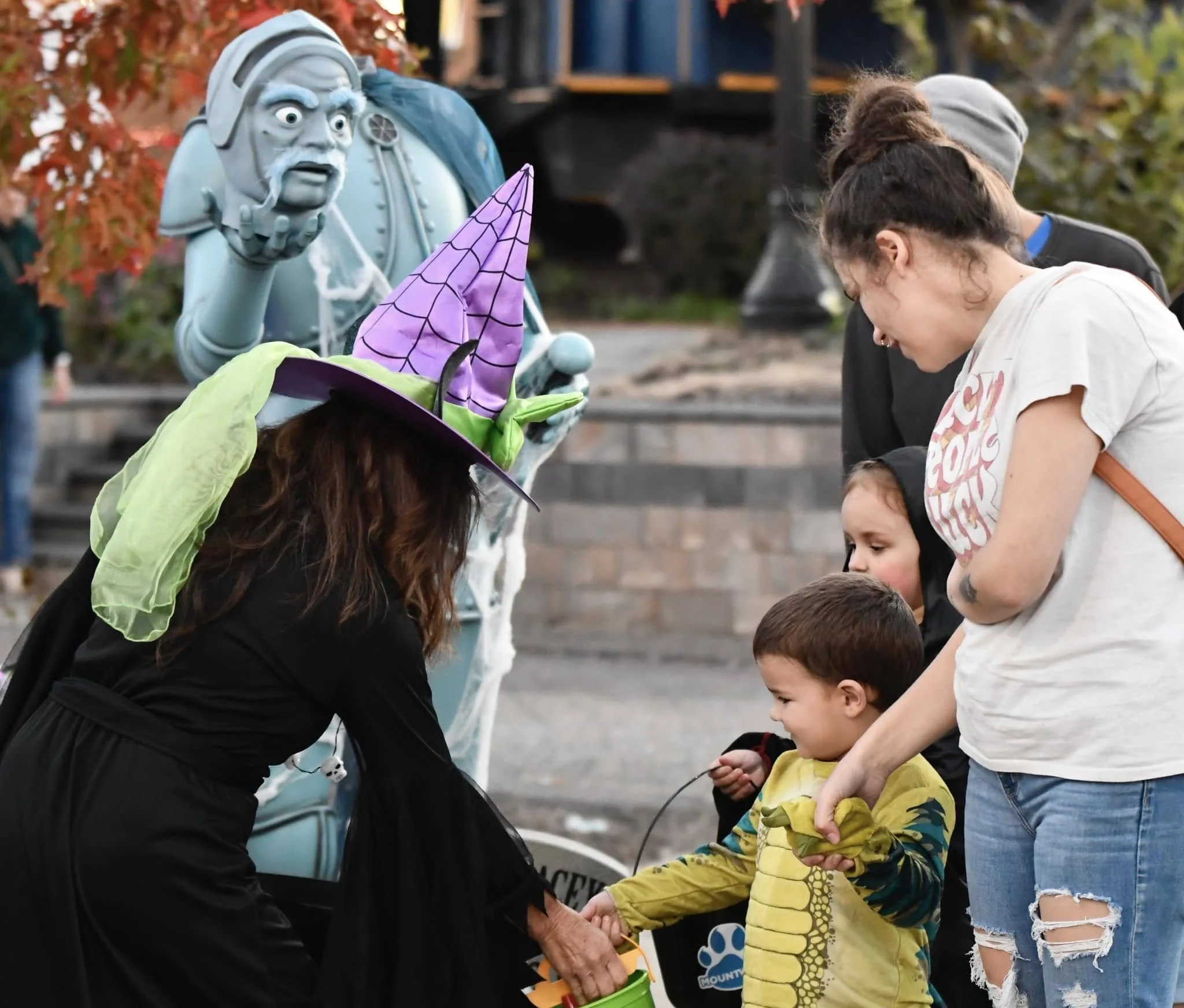 A woman dressed as a witch handing out sweets to children in a Halloween costume parade, with a witch and skeleton decoration in the background.
