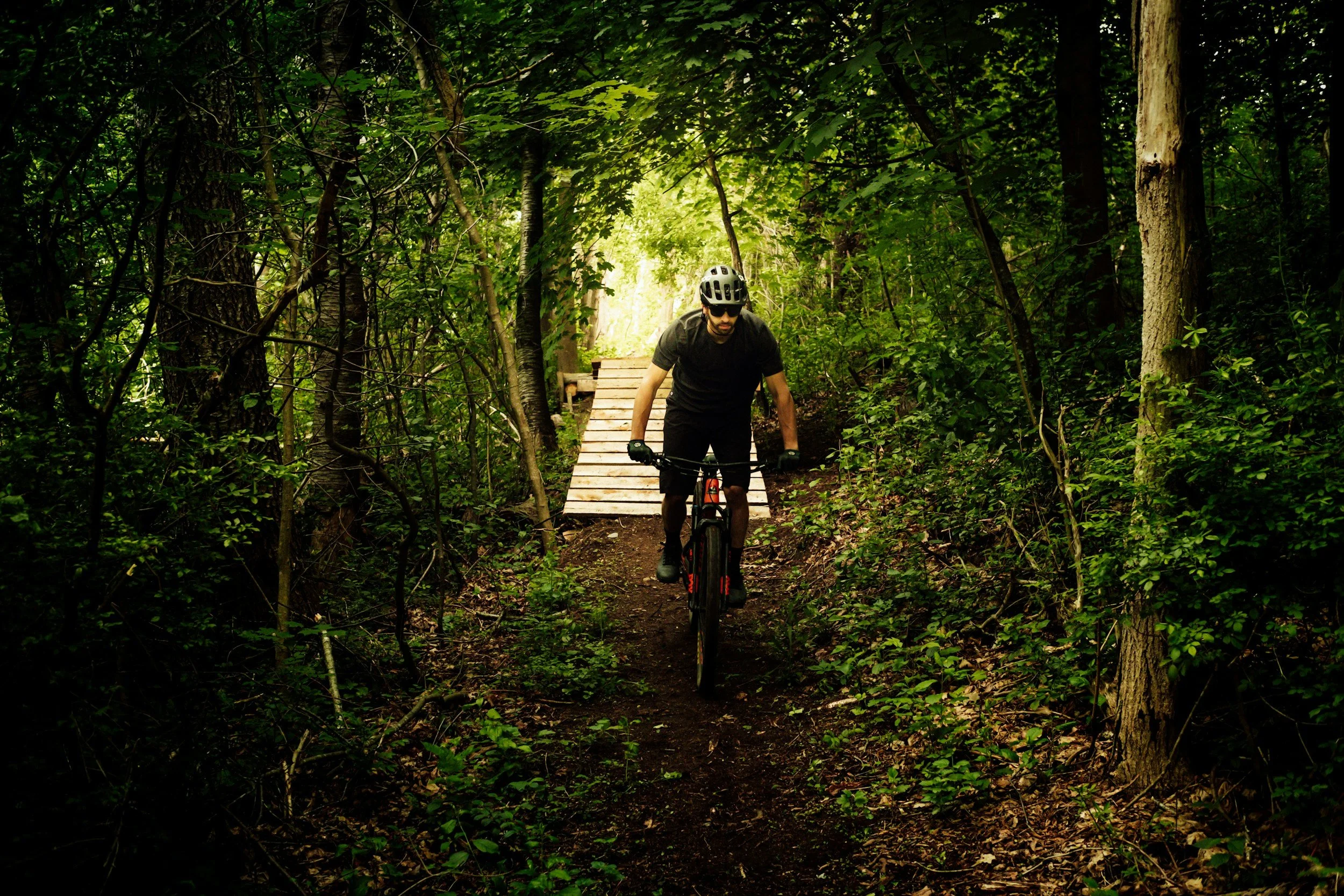 A man wearing a helmet and sunglasses biking on a forest trail, with a wooden bridge ahead.