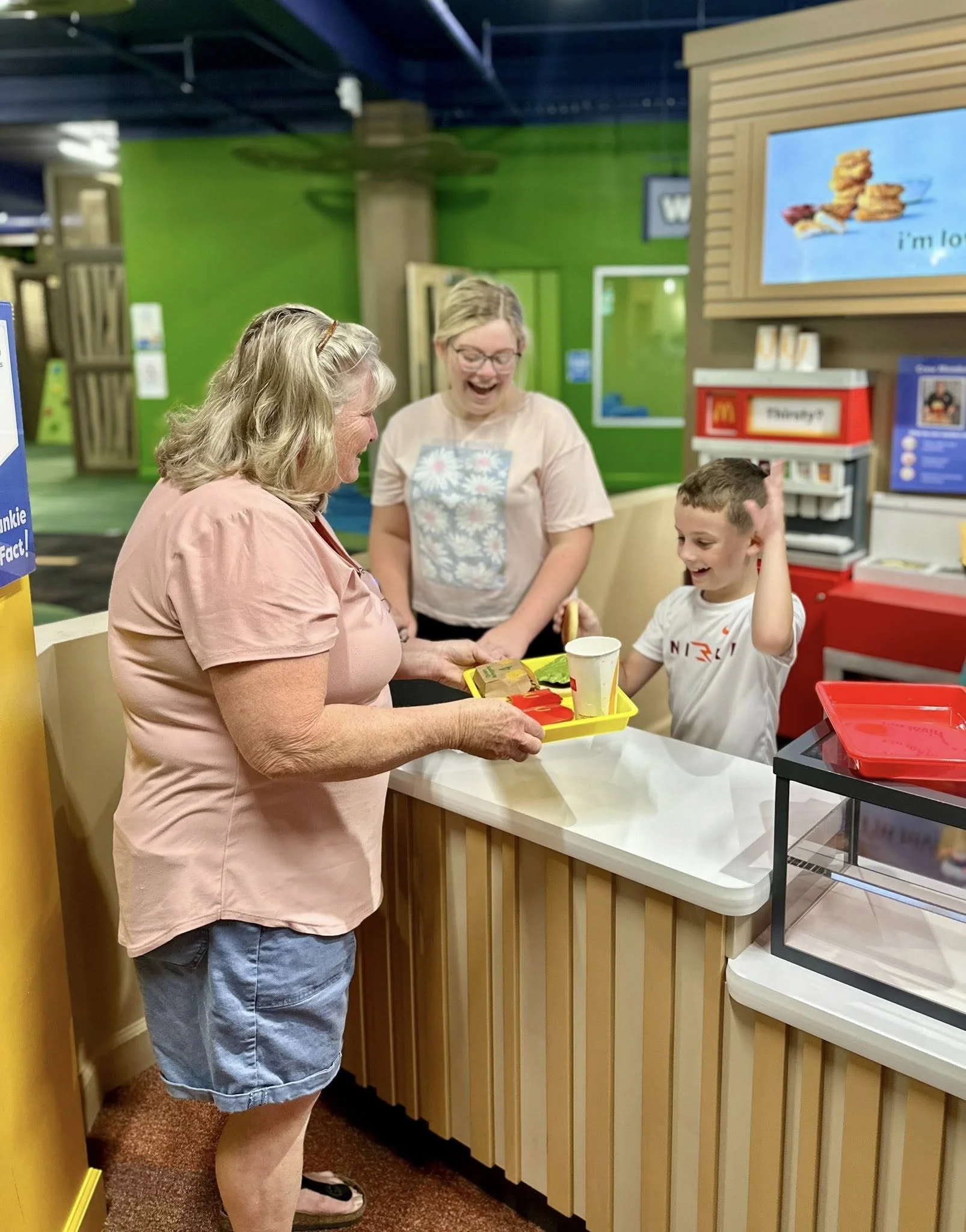 A woman and a young boy are at the counter of a fast food restaurant, with another woman behind them. The woman is holding a yellow tray with food, and the boy is smiling with one hand on his head, wearing a white shirt. The background features a green wall and a digital display showing food items.
