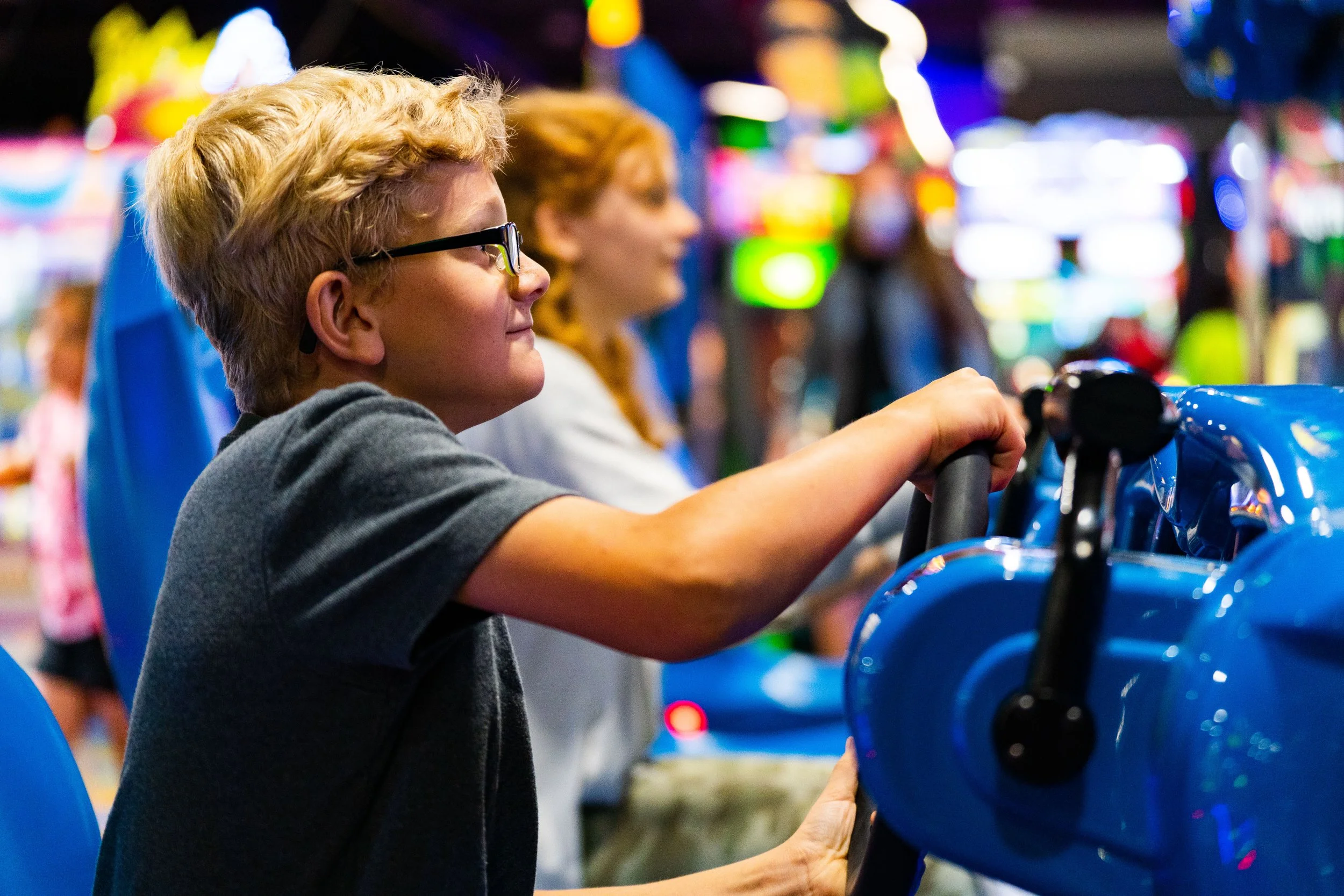 A young boy with glasses playing an arcade racing game in an amusement arcade, with colorful blurred arcade lights and other kids in the background.