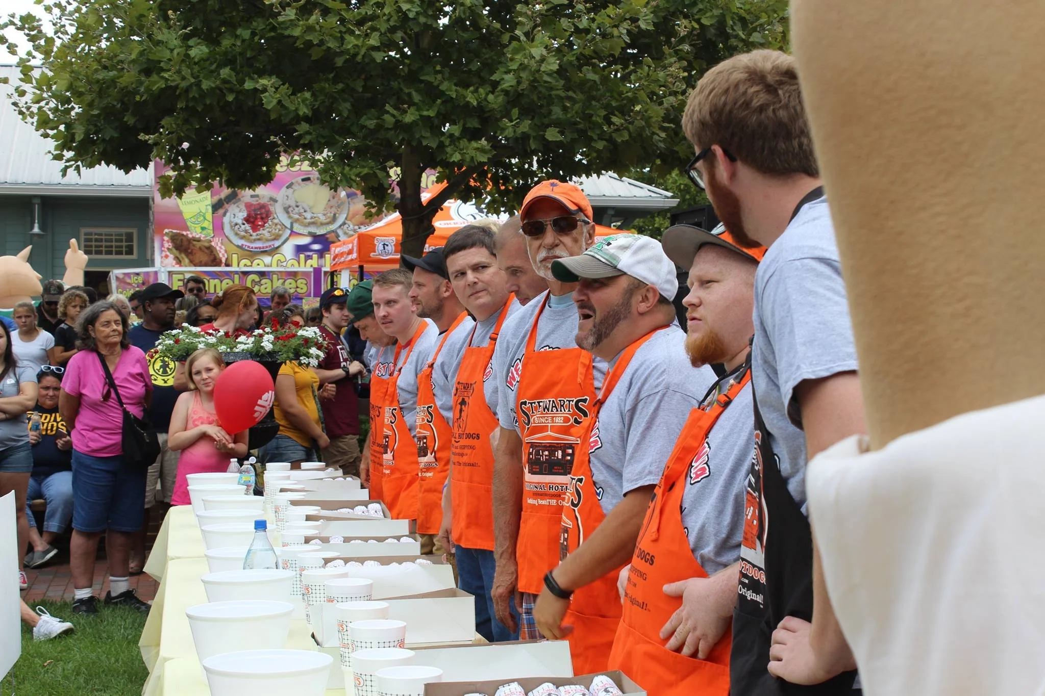 A group of men wearing gray shirts and orange aprons standing in a line at an outdoor event, with spectators watching. There are tables with bowls and items in front of them, and a crowd in the background near food stalls.