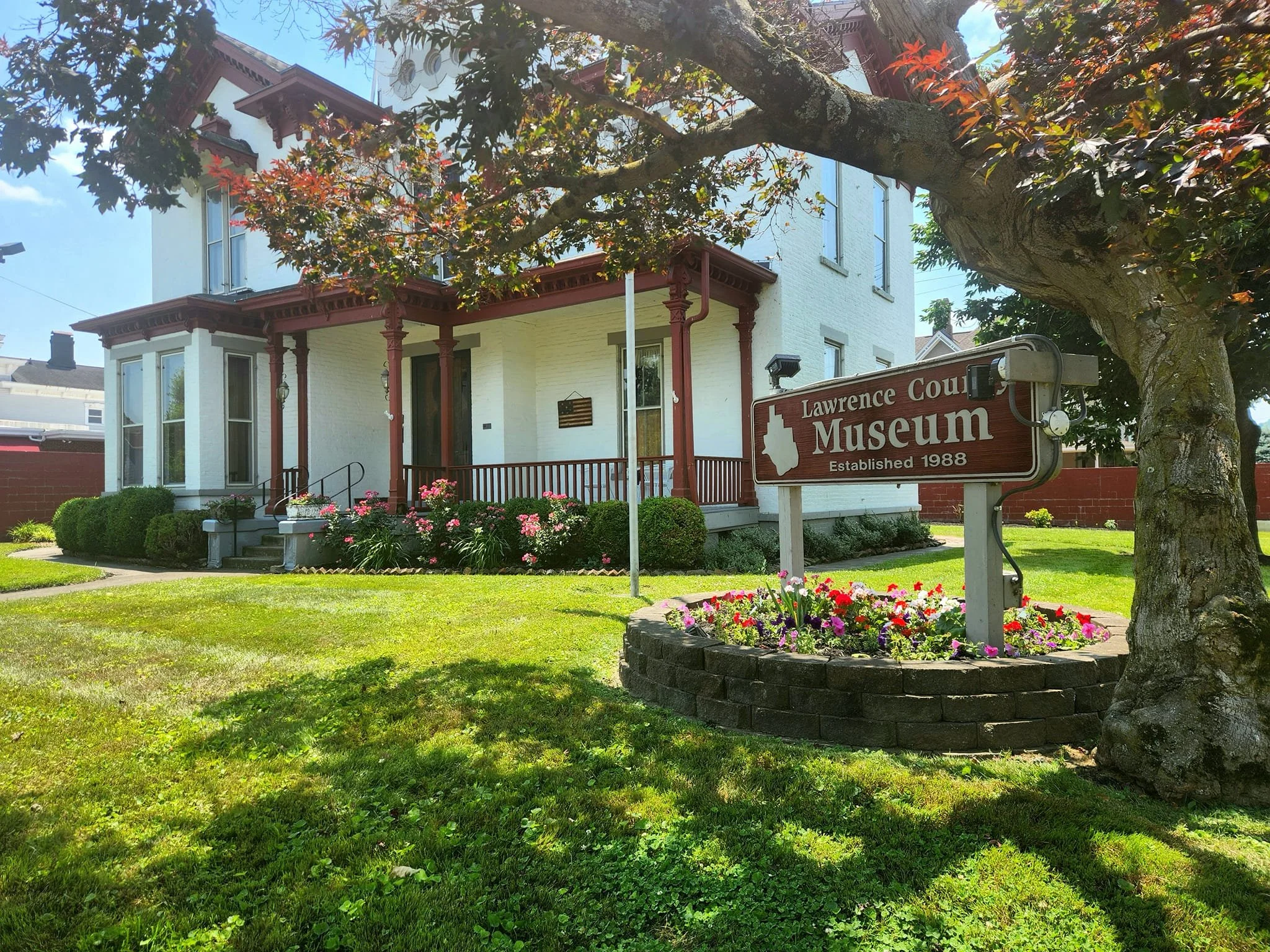 The image shows a white historic house with a front porch, surrounded by a well-kept garden with colorful flowers, and a sign indicating it is the Lawrence County Museum established in 1988, located next to a large tree.