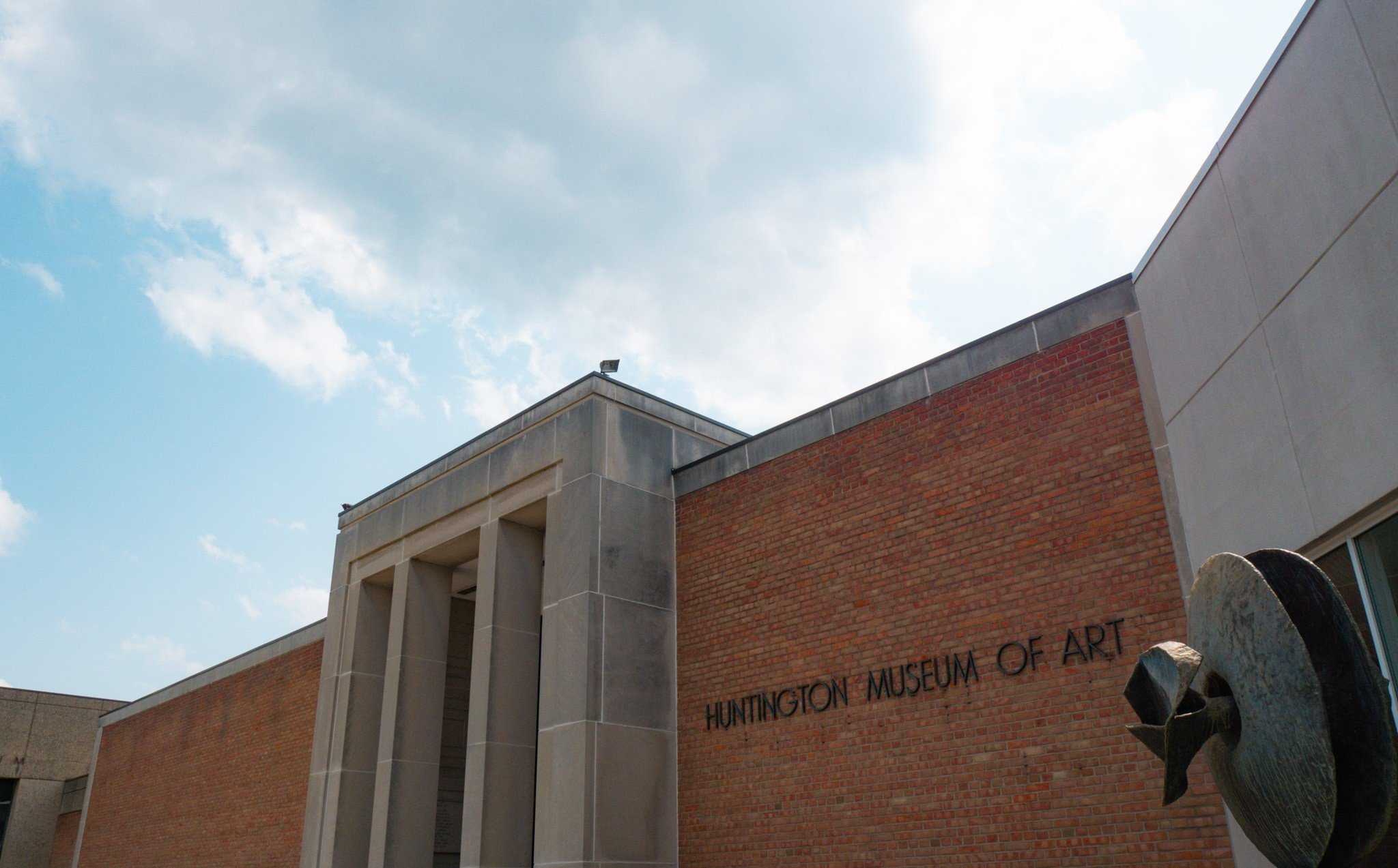 Exterior of the Huntington Museum of Art building with modern architectural design, brick and concrete walls, and a sculpture visible in the foreground, under a partly cloudy sky.