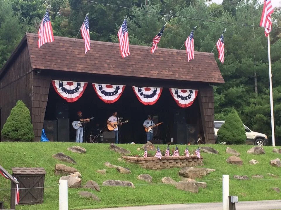 A small outdoor stage decorated with American flags and patriotic bunting, with a band performing on a grassy hillside, surrounded by rocks and green trees.