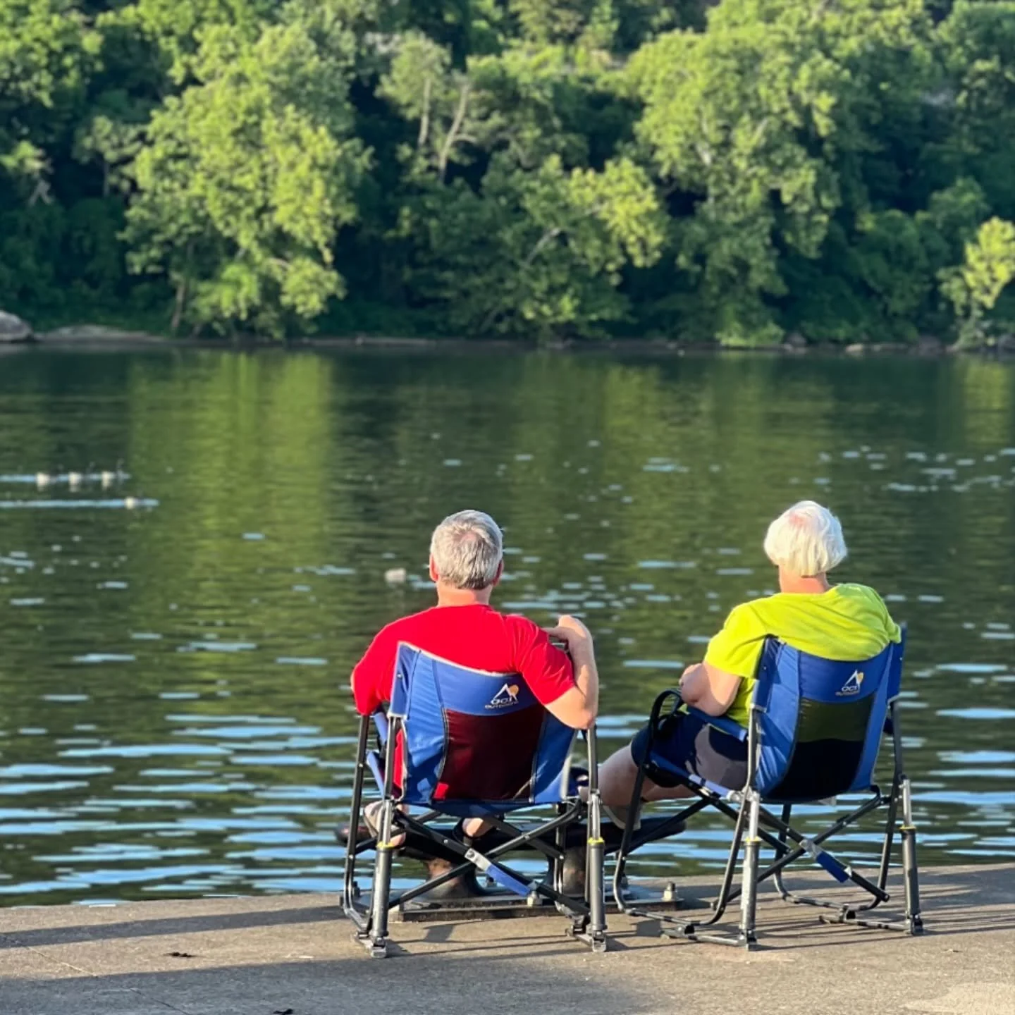 Two elderly people sitting in camping chairs by a lake, facing the water, with a tree-lined shore in the background.