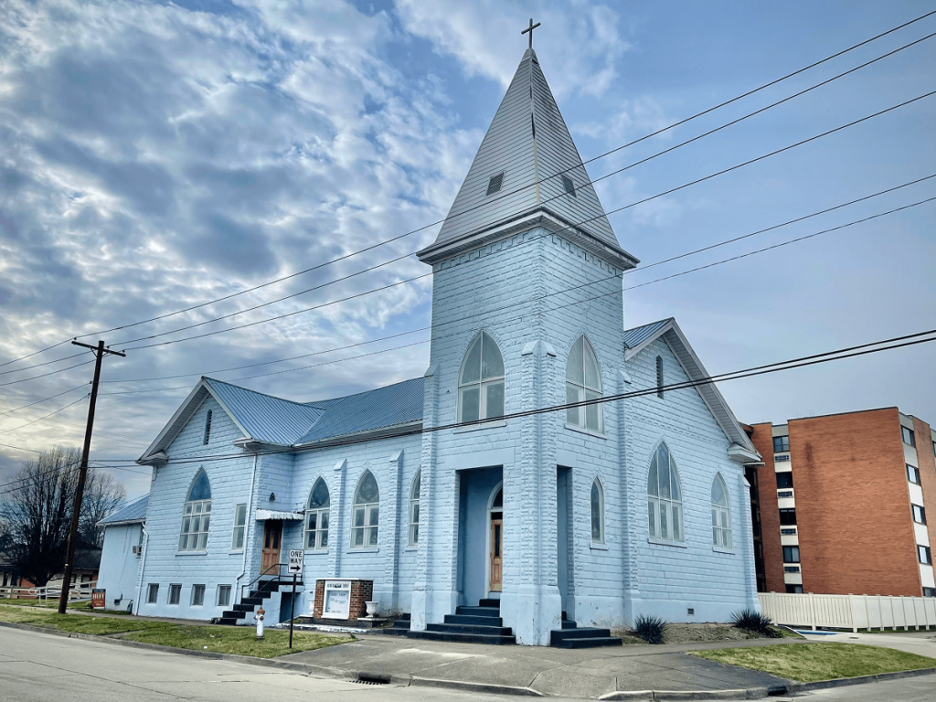 White church building with a tall steeple and multiple pointed arched windows, situated on a street with power lines and a parking area.