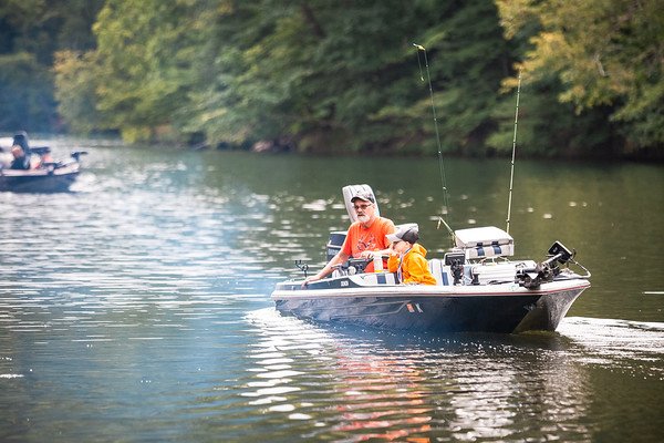 An older man wearing a white hat and orange shirt fishing on a small boat in a river surrounded by green trees.