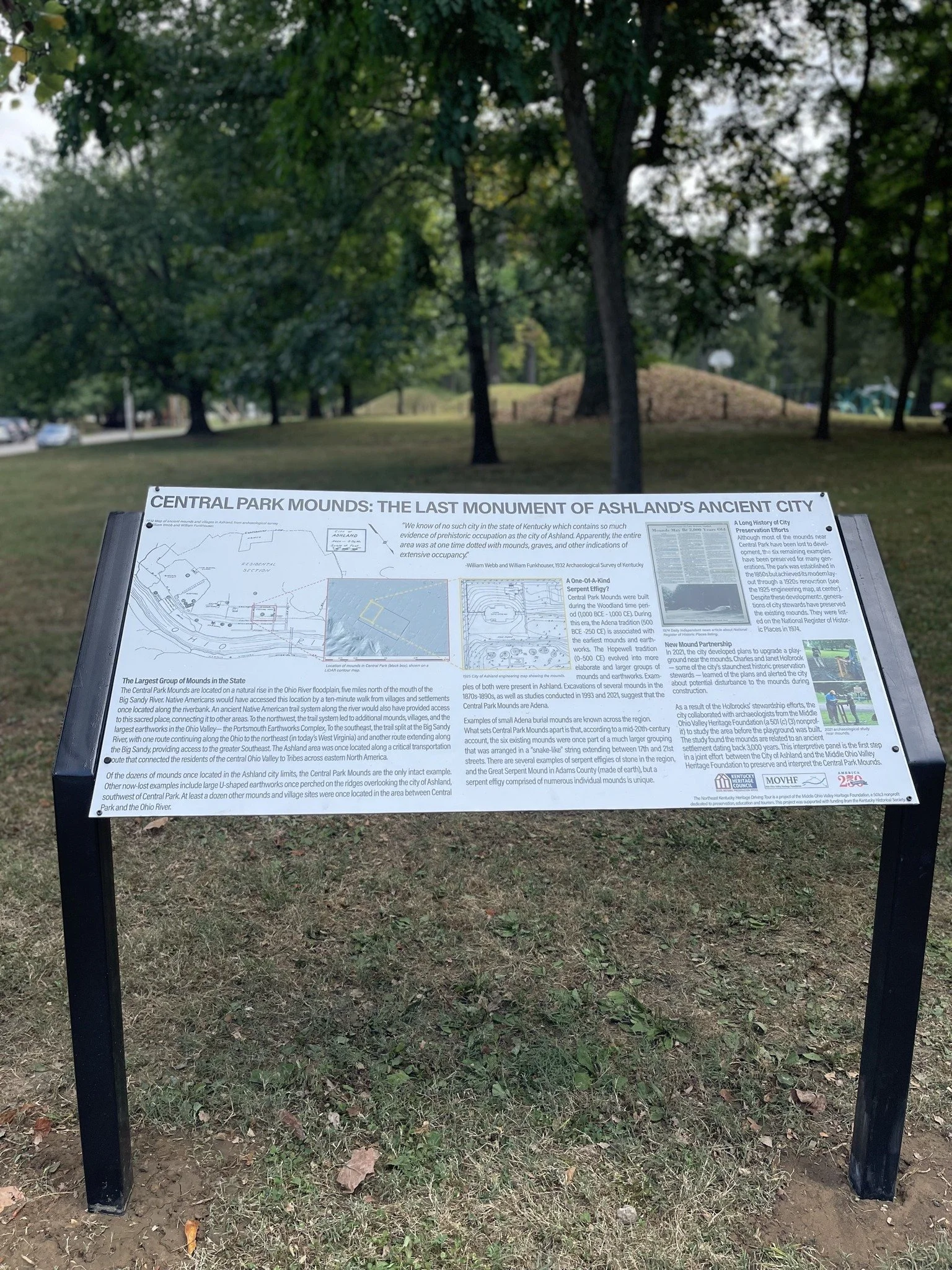 An informational sign titled 'Central Park Mounds: The Last Monument of Ashland's Ancient City' in a park setting with trees and grass in the background.