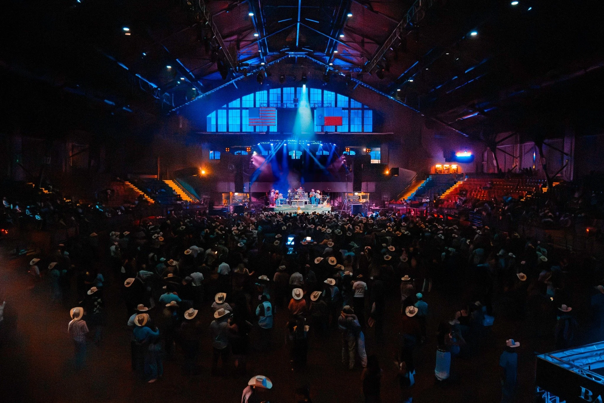 A large concert hall filled with a crowd of people watching a live music performance on stage. The stage is illuminated with blue and purple lights, and the audience is wearing cowboy hats.
