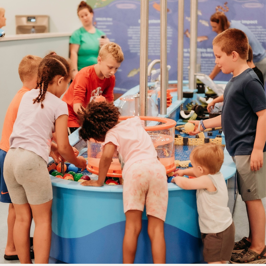 Children playing with water toys at an indoor water-play area, supervised by adults in the background.