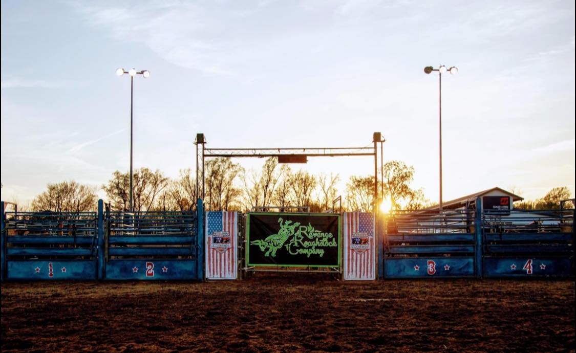 Empty rodeo arena with gates labeled 1 to 4, a digital sign with a green dragon logo, and trees in the background during sunset.