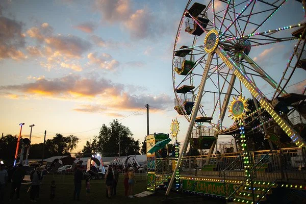A carnival scene at sunset with a large Ferris wheel lit with colorful lights, and people walking around on the grassy area.