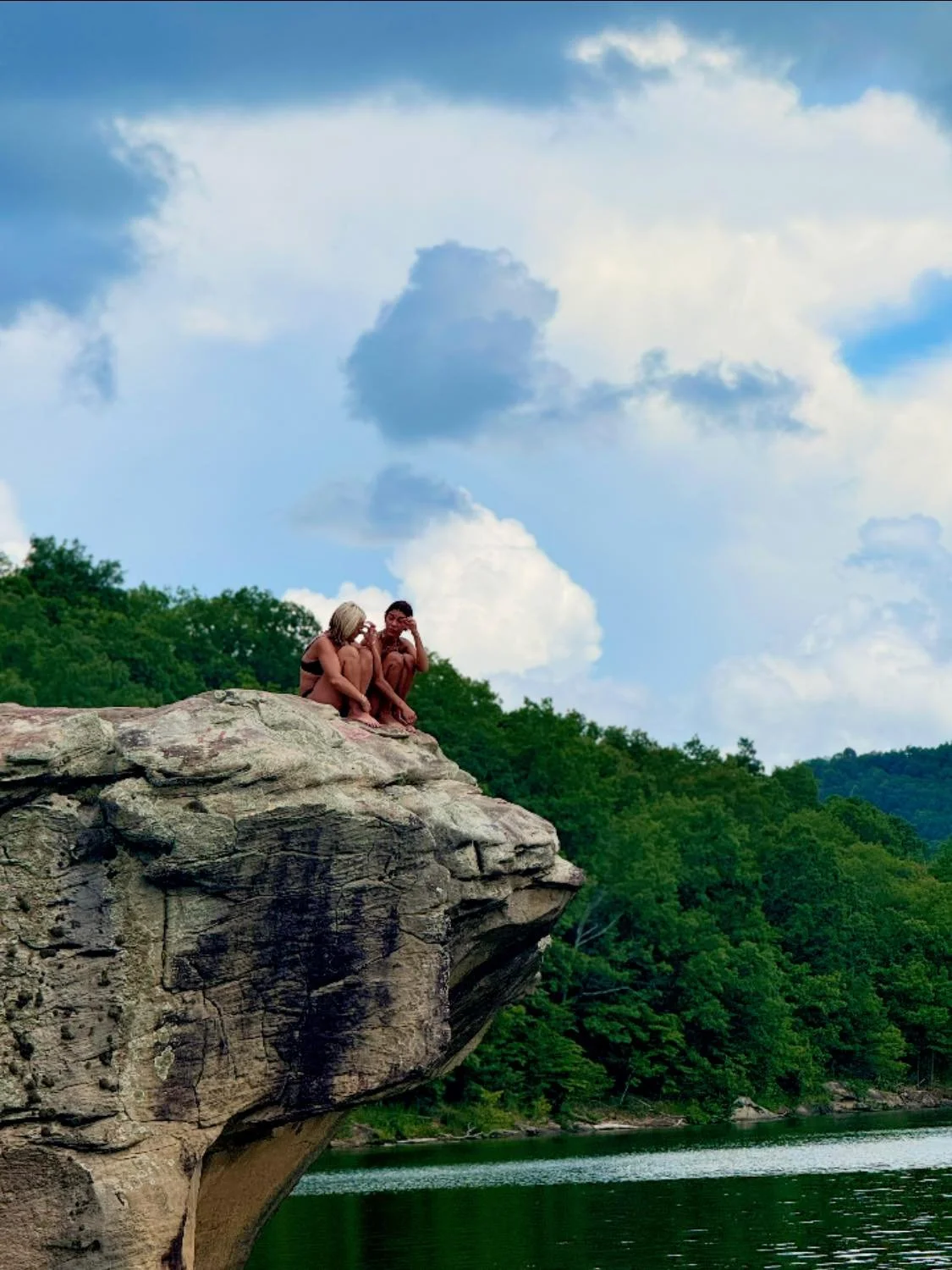 Two children sitting on a rocky ledge overlooking a river, surrounded by lush green trees and a cloudy sky.