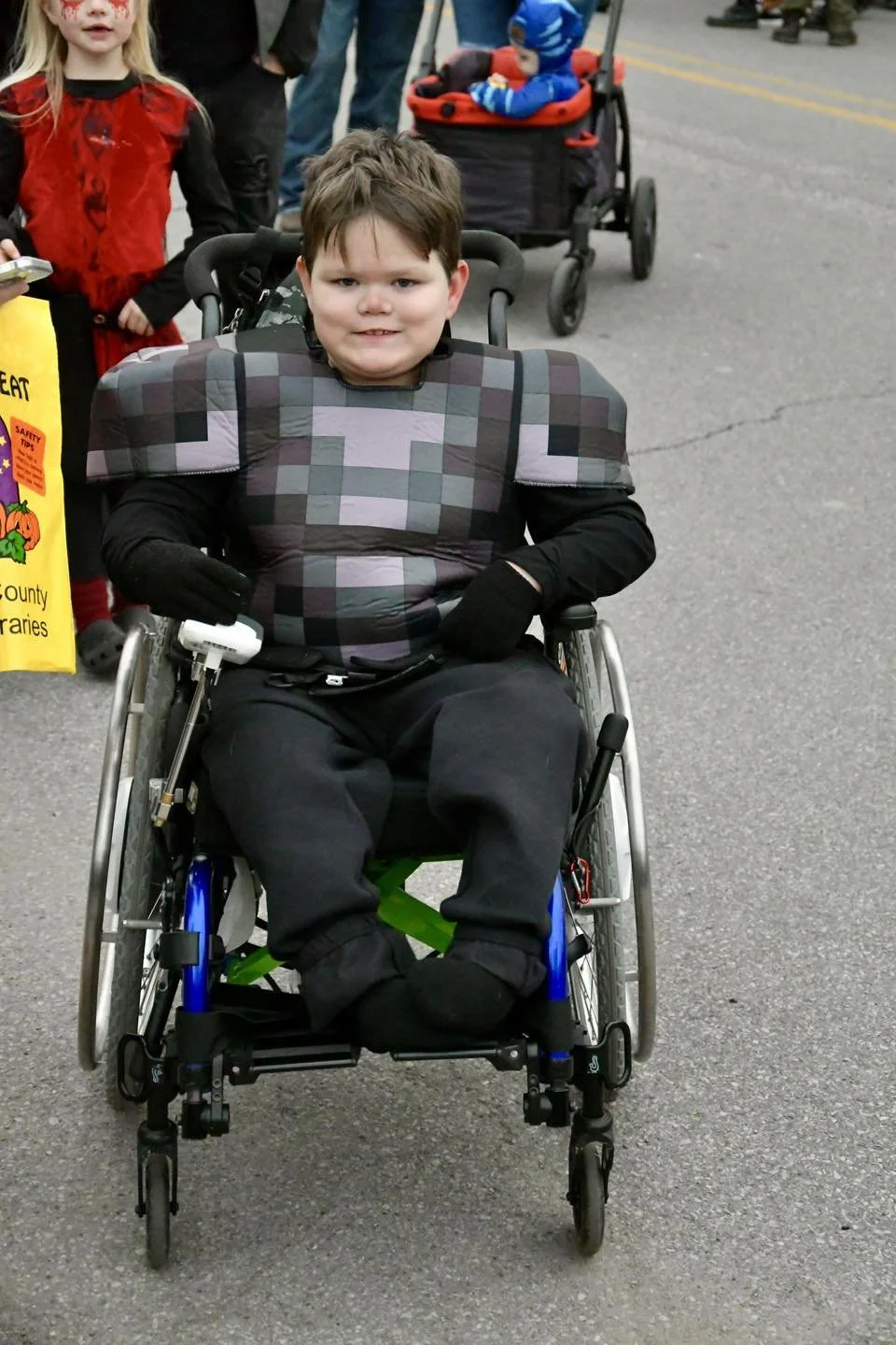 A young boy in a wheelchair wearing a pixelated patterned shirt, black pants, and gloves. Behind him, a girl dressed as a ladybug and a baby in a stroller, with other people in the background on a paved street.