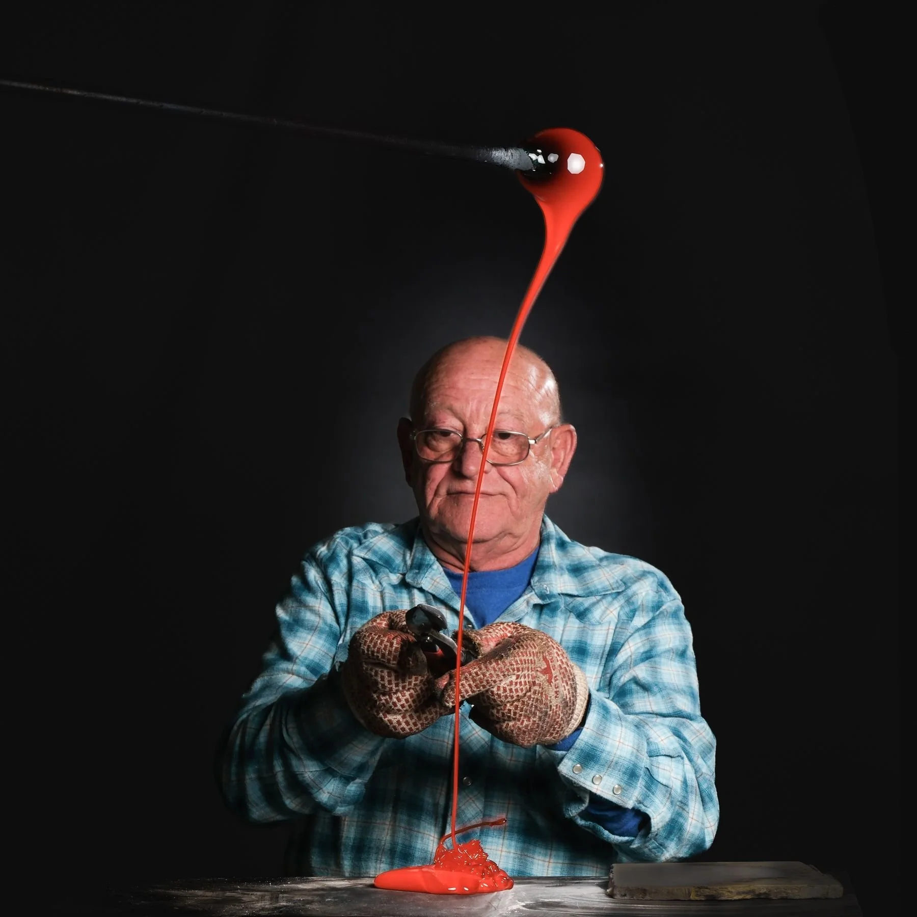 An elderly man wearing glasses, a blue plaid shirt, and gloves holding a black pair of pliers, with bright red liquid pouring from a spoon above his head onto the table, creating a puddle.