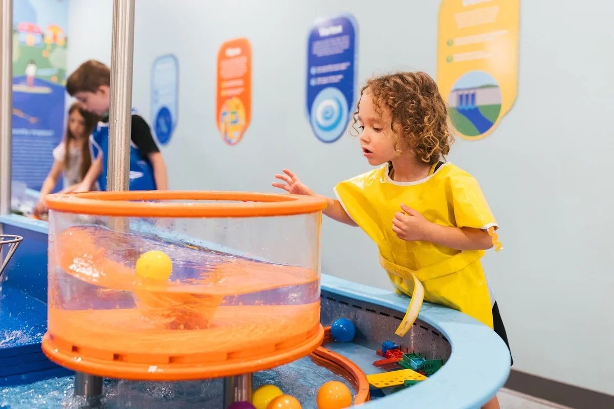 A young girl wearing a yellow smock plays at a water table with a bubble tube and floating balls, in an interactive exhibit area with informational posters on the wall in the background.