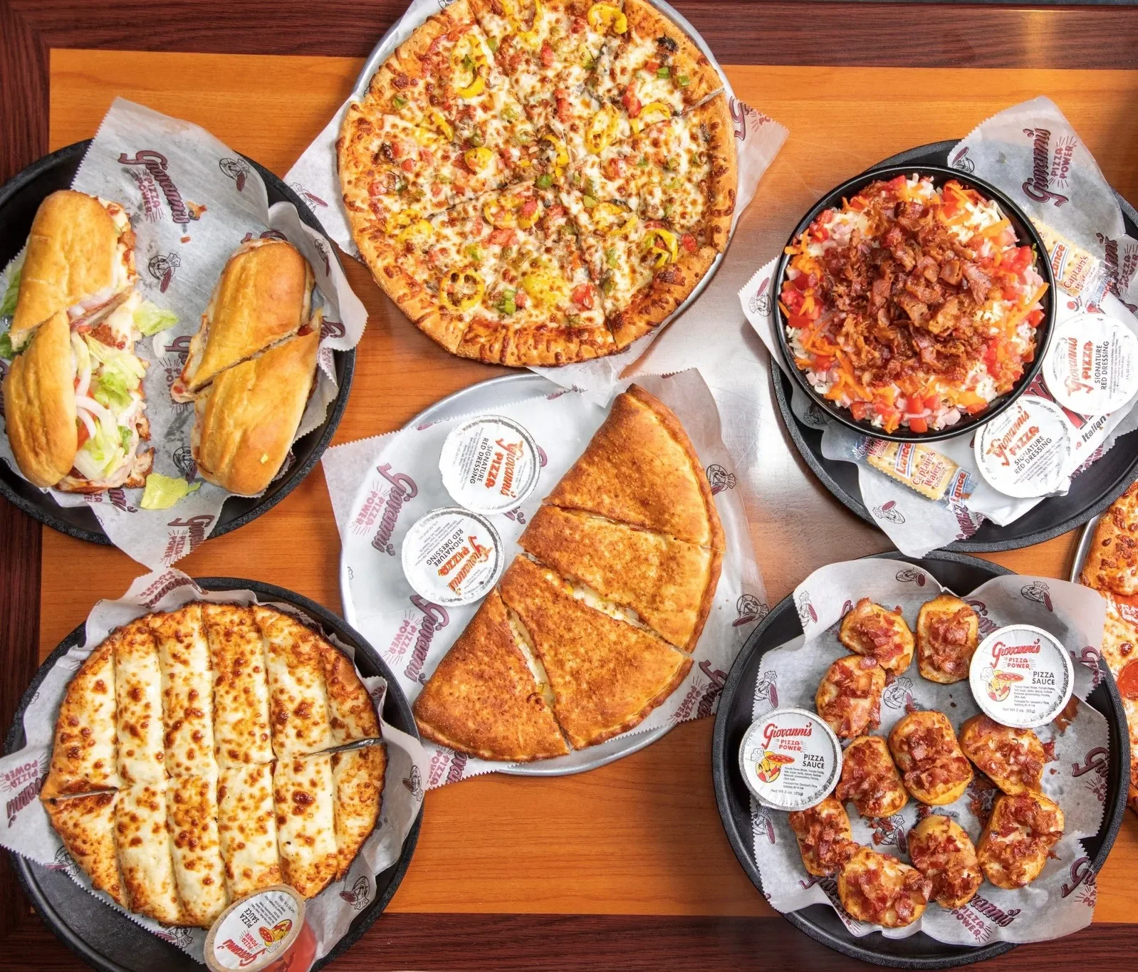 Assorted Italian-style dishes on a wooden table, including two sandwiches, two pizzas, a baked cheese dish, a sub, and a plate of mini pepperoni pizzas, all served with pizza sauce packets.