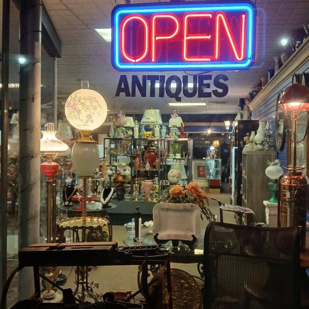 The image shows the interior of an antique store with various vintage lamps, vases, and decor items. A bright neon sign above the window reads 'OPEN' in red and blue colors, and below it, a sign says 'ANTIQUES.' The shop is filled with classic and vintage items.