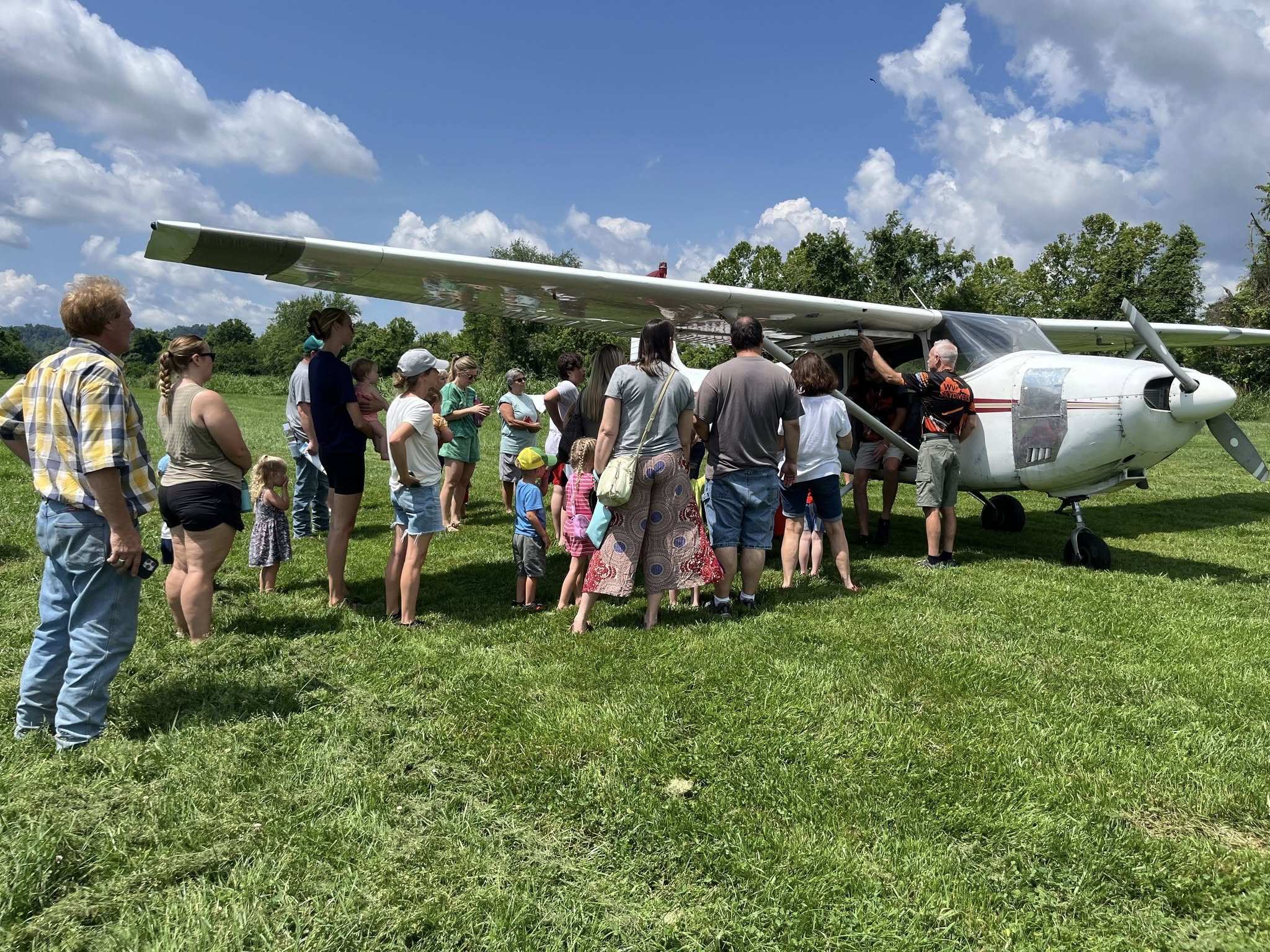 Group of people, including children and adults, gathered around a small aircraft on a grassy field on a sunny day.