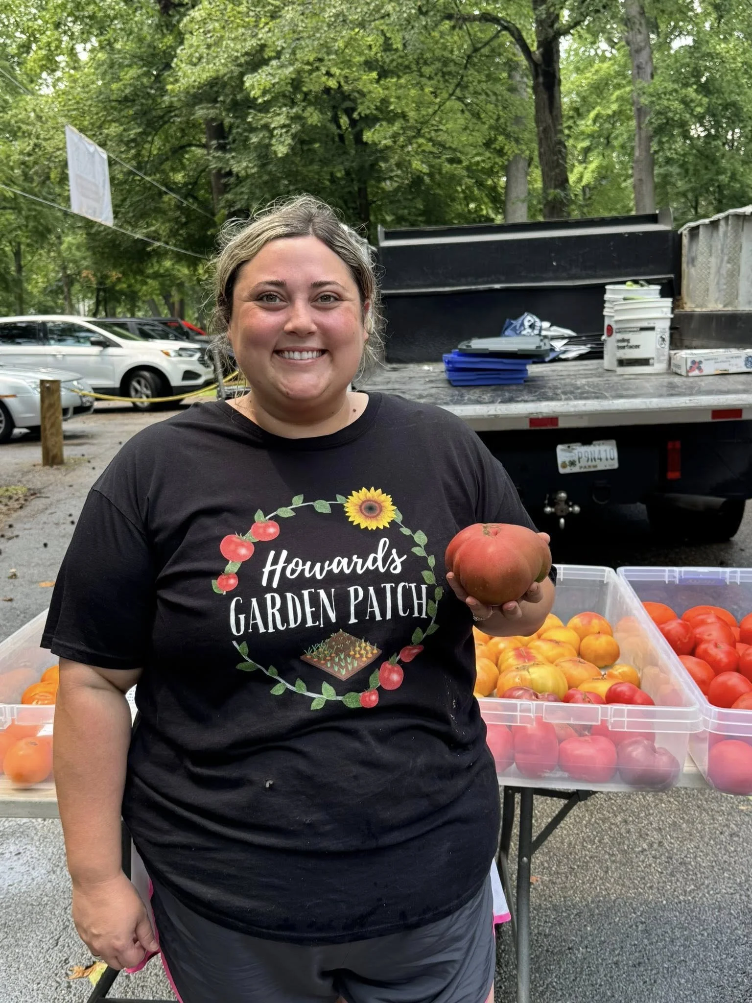 A woman smiling and holding a large tomato at a farmers market stand with trays of tomatoes behind her, wearing a black t-shirt with 'Howards Garden Patch' printed on it.
