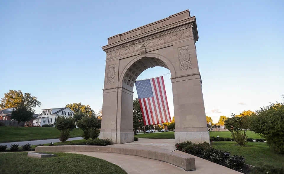 Stone arch monument with an American flag hanging through it, surrounded by grass, trees, and houses in the background.