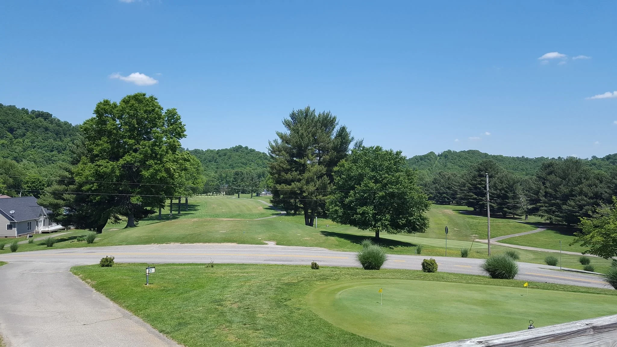 A scenic view of a golf course with green grass, a putting green with flags, several trees, a road, utility poles, and distant hills under a clear blue sky.