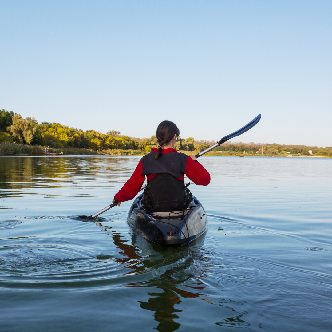 Person kayaking on a calm river during daytime with clear skies and green trees in the background.