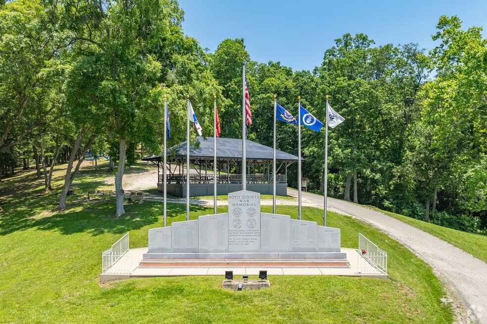 Boyd County war memorial with a stone plaque, surrounded by eight flags, and a pavilion in the background, located in a green park with trees and a gravel path.