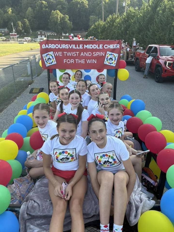 Group of young girls in white t-shirts riding on a decorated float at a parade, with colorful balloons and a banner that reads 'Barboursville Middle Cheer, We Twist and Spin for the Win'.